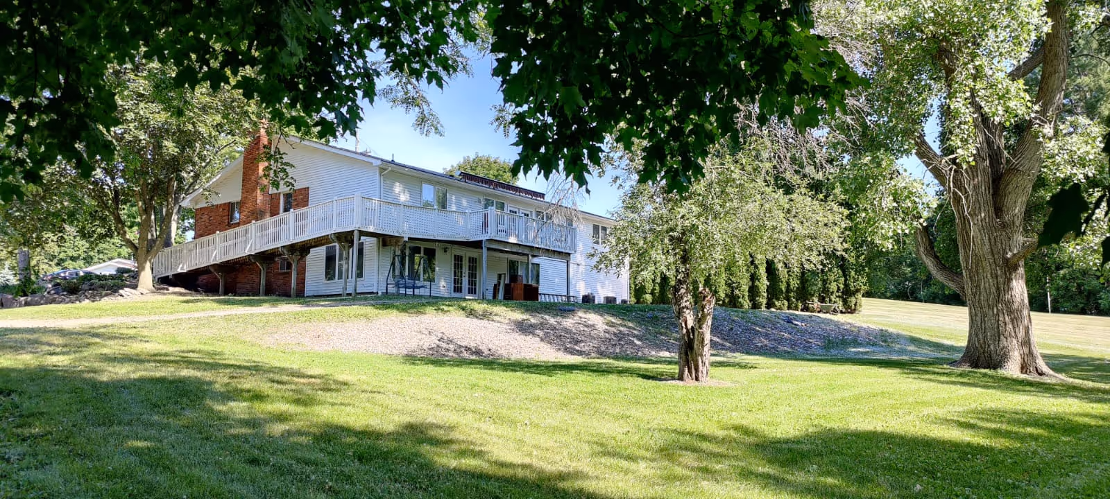 A two-story white building with a wraparound balcony on the upper floor, surrounded by green grass and large trees under a clear blue sky.