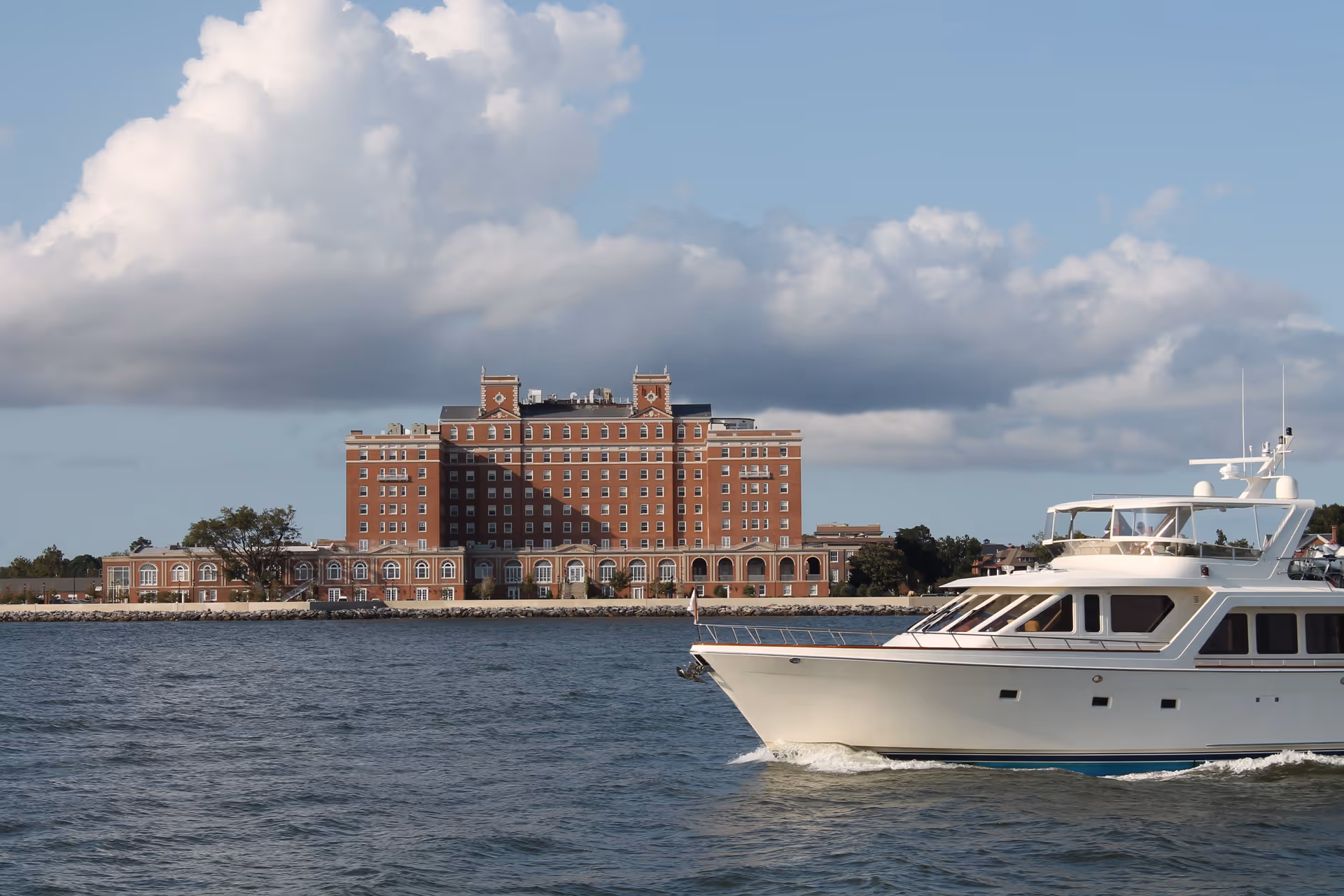 A large brick building with multiple windows and two small towers on the roof, situated along a waterfront with a boat cruising in the foreground under a partly cloudy sky.
