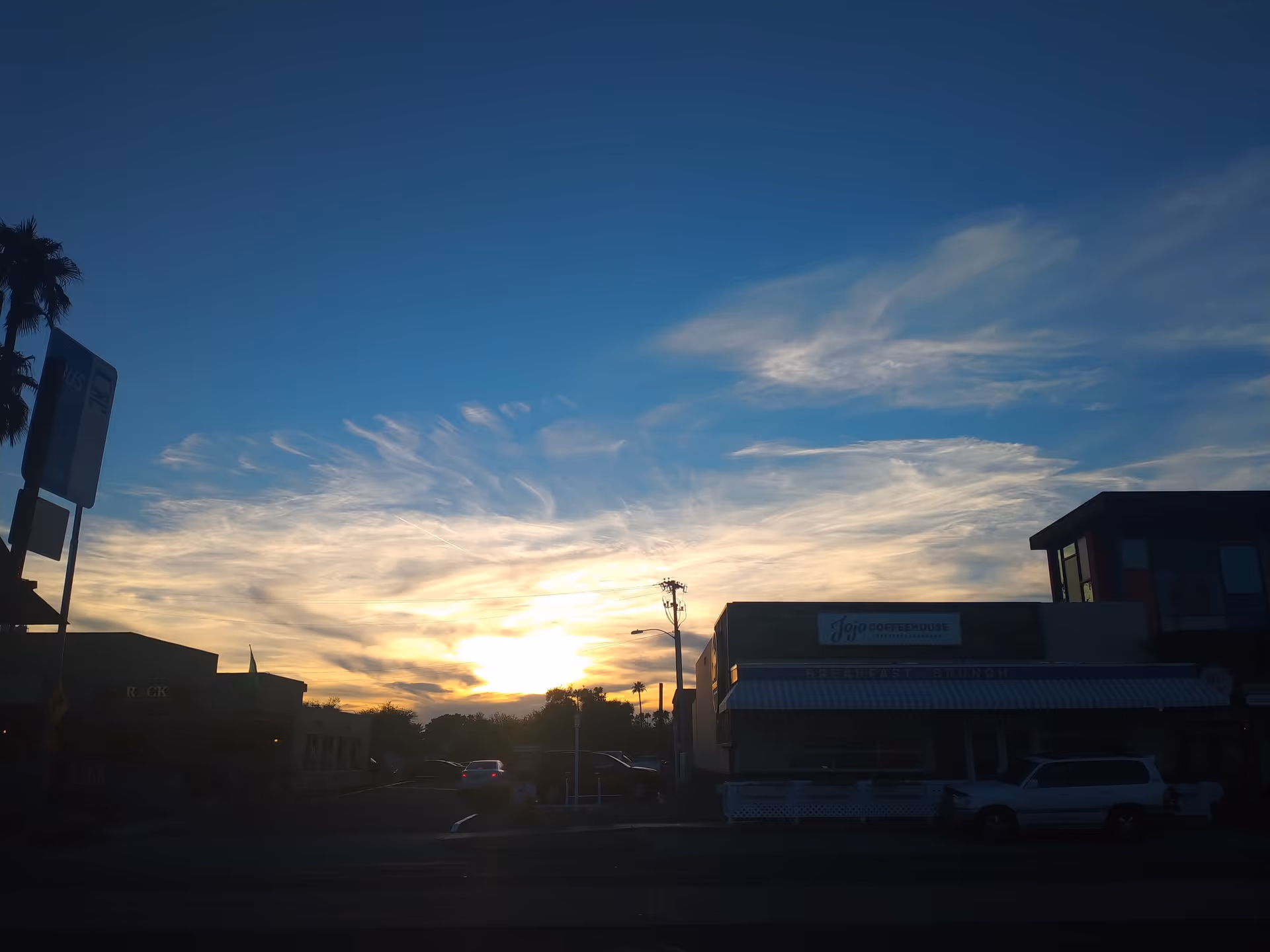 Sunset over a street with storefronts, parked cars, and a blue sky.