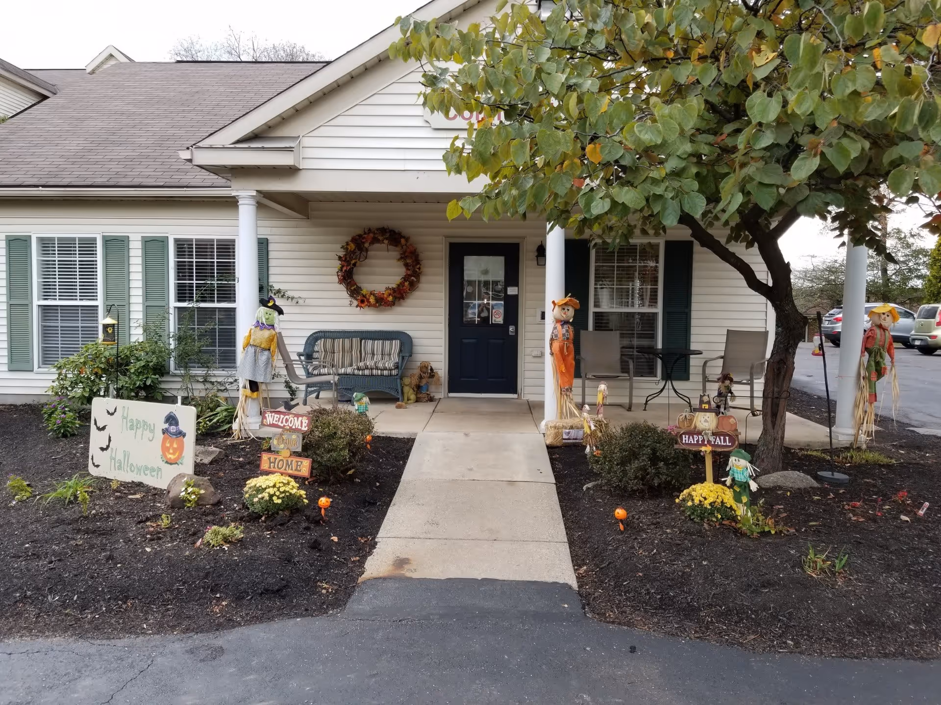Front entrance of a single-story assisted living facility decorated with Halloween and fall-themed decorations including scarecrows, a wreath on the wall, and signs that say 'Happy Halloween', 'Welcome Home', and 'Happy Fall'. There is a bench and two chairs with a small table on the porch, and a tree with green leaves on the right side.