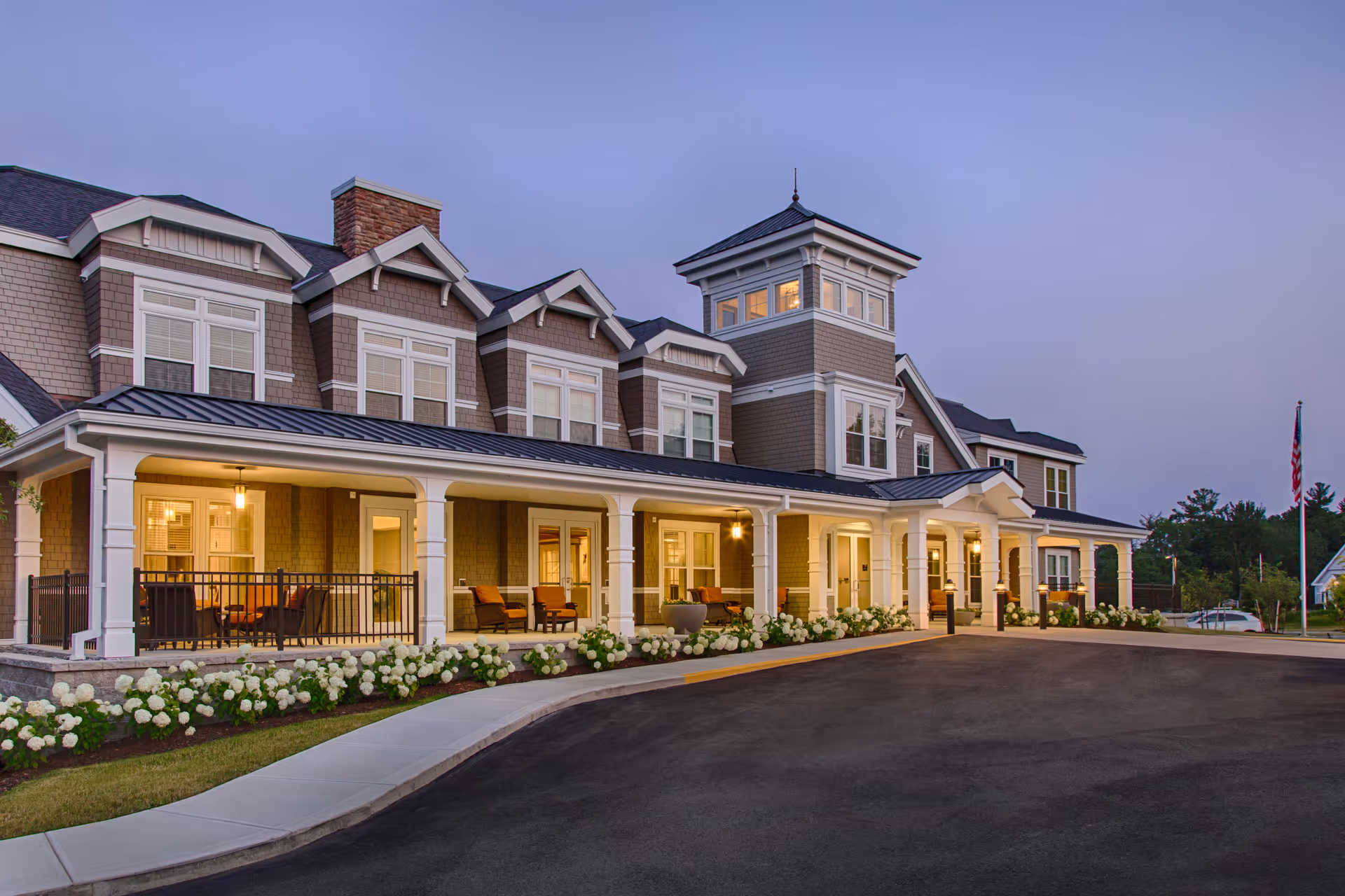 Exterior view of a large, elegant senior living facility building at dusk with warm lights glowing from inside. The building features multiple windows, a covered porch with seating, white columns, and a landscaped area with white flowers along the sidewalk and driveway.