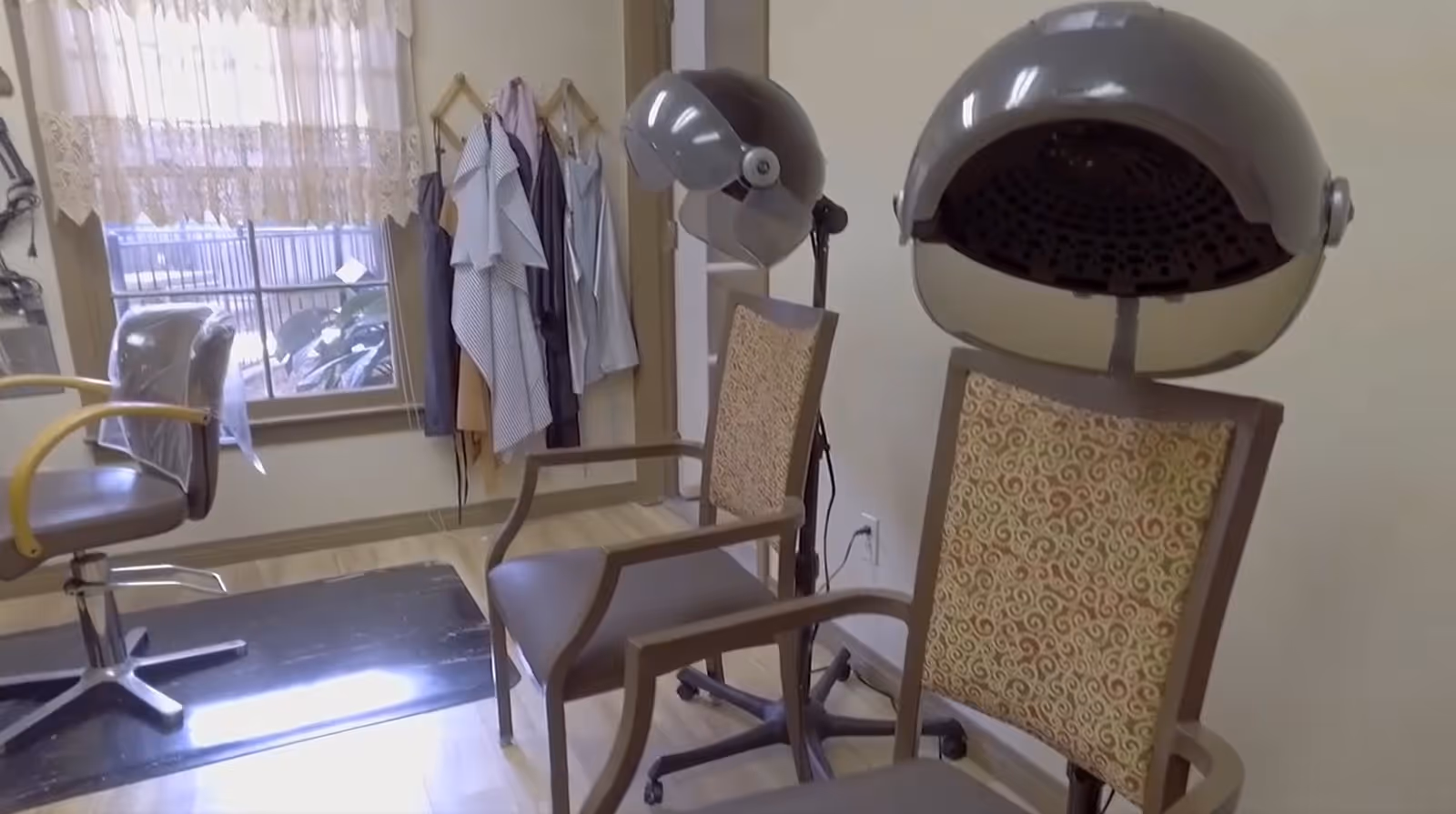 Interior view of a hair salon area in Lafreniere Assisted Living Center featuring two vintage hair drying chairs with hooded dryers, a salon chair, a window with lace curtains, and several hanging capes or towels on the wall.