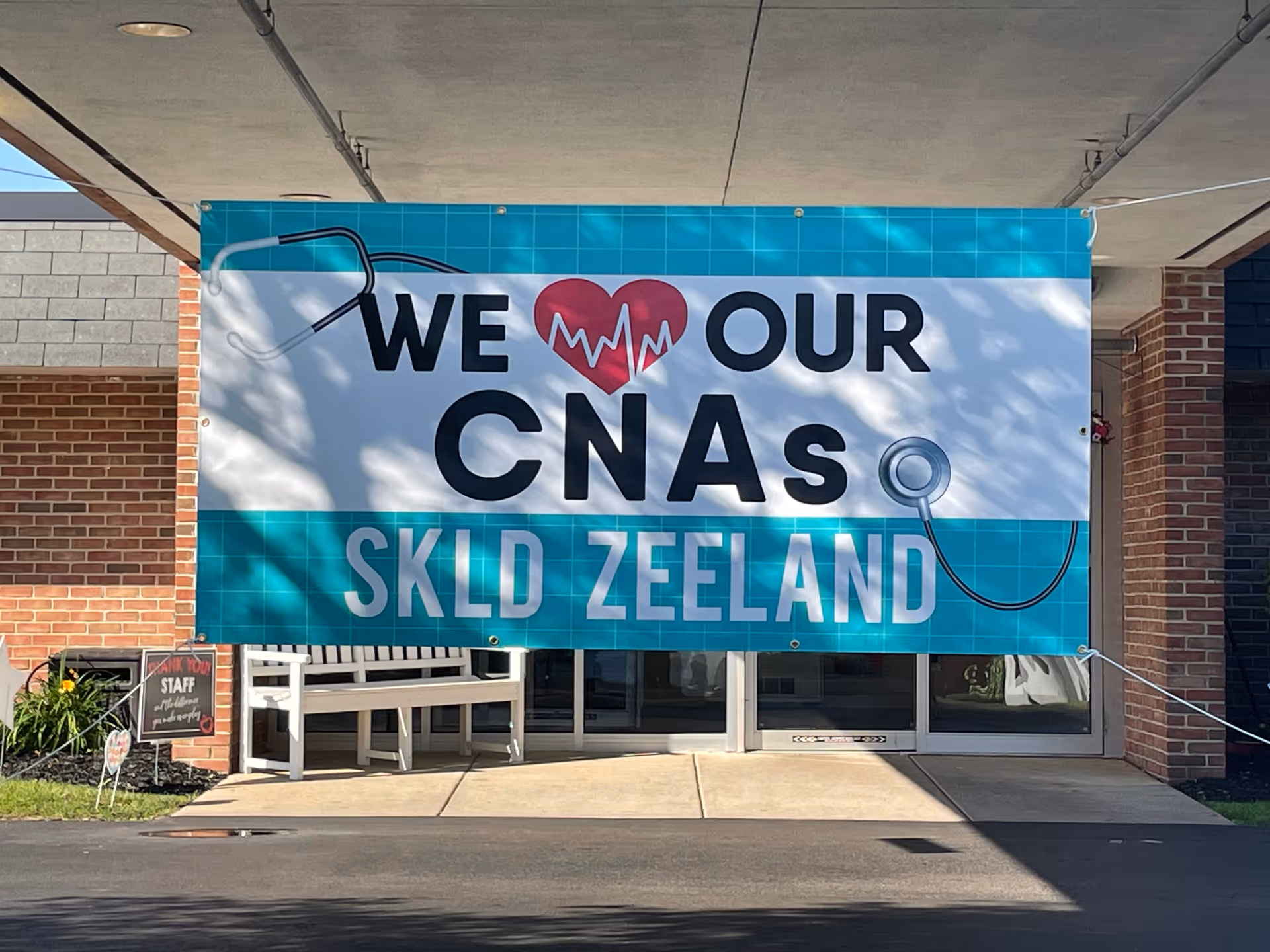 A large banner hanging outside a building entrance reads 'We love our CNAs SKLD Zeeland' with a heart symbol and a stethoscope graphic on a blue and white background.
