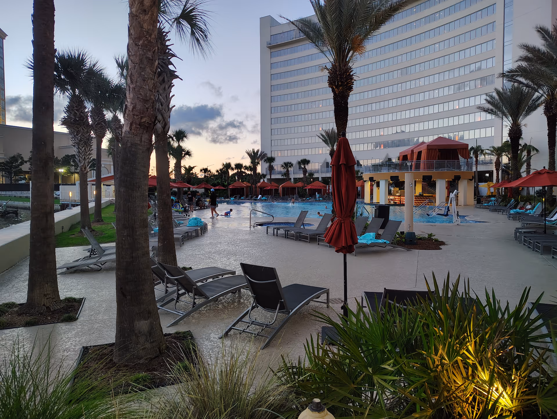 Outdoor pool area with lounge chairs, red umbrellas, palm trees, and a tall building.