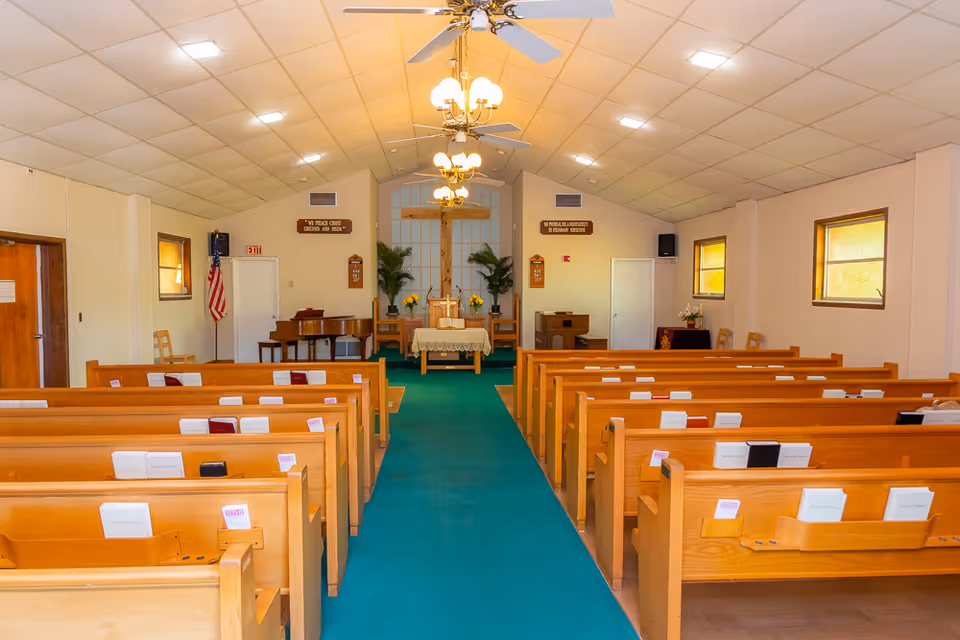 Interior view of a chapel with wooden pews arranged in rows on either side of a green carpeted aisle leading to an altar with a cross, flowers, and a piano in the background. The ceiling has white tiles with ceiling fans and chandeliers, and there are windows on the right wall letting in natural light.