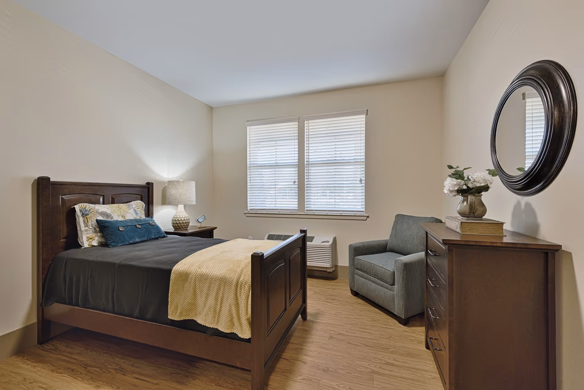 Small furnished bedroom with a wooden bed, nightstand and lamp, armchair, dresser topped with flowers and round wall mirror, and a window with blinds.