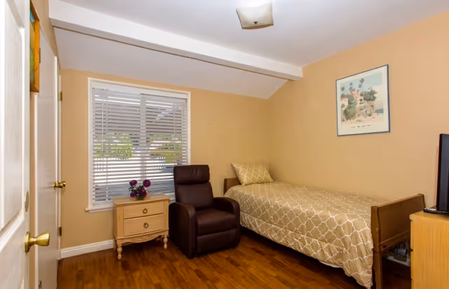 A modest bedroom in a senior living facility with a bed, chair, and dresser.