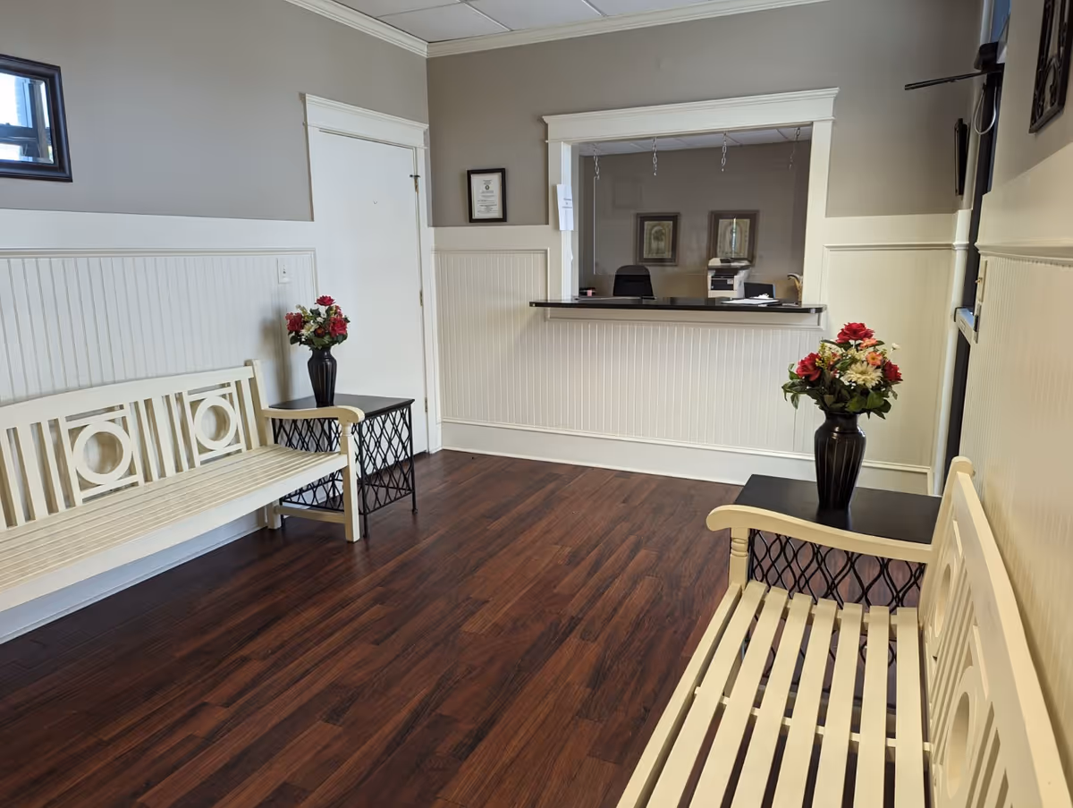 Reception area with white benches, side tables, flower vases, and a front desk window.