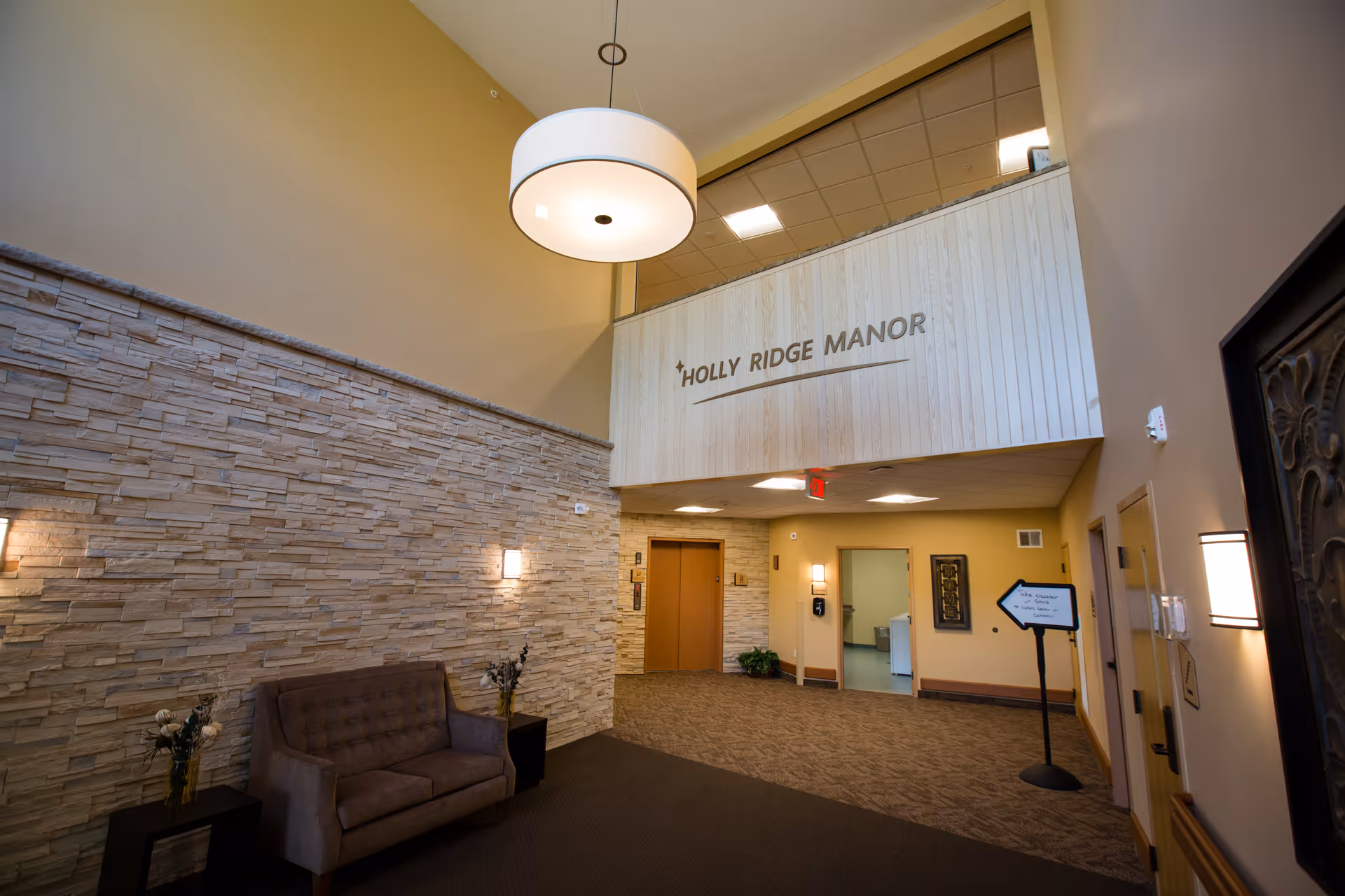 Lobby interior of Holly Ridge Manor showing a stone accent wall, tufted sofa, overhead pendant light, and a mezzanine sign reading 'HOLLY RIDGE MANOR'.