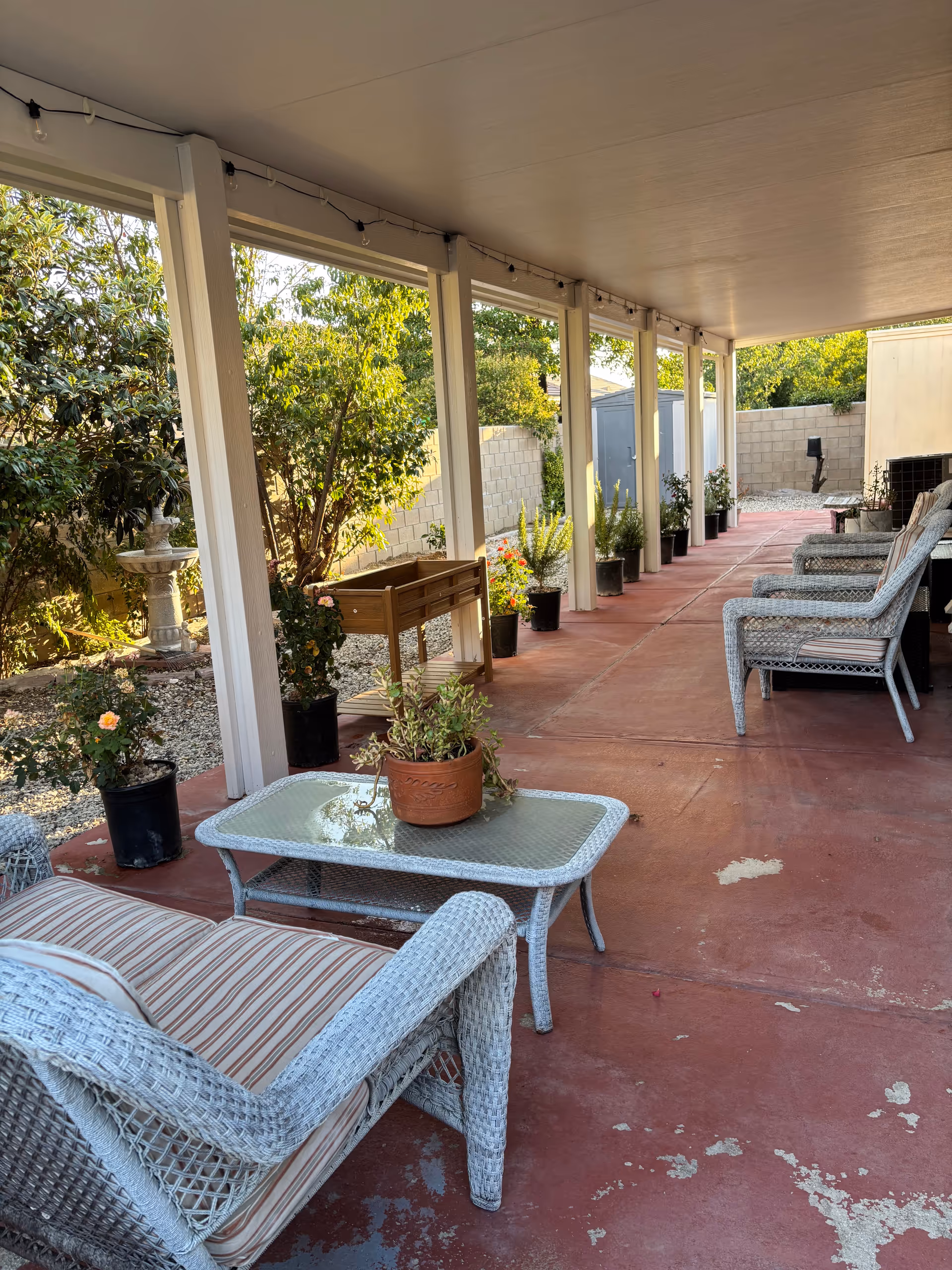 Covered outdoor patio with wicker chairs, glass tables, and potted plants along a walkway.