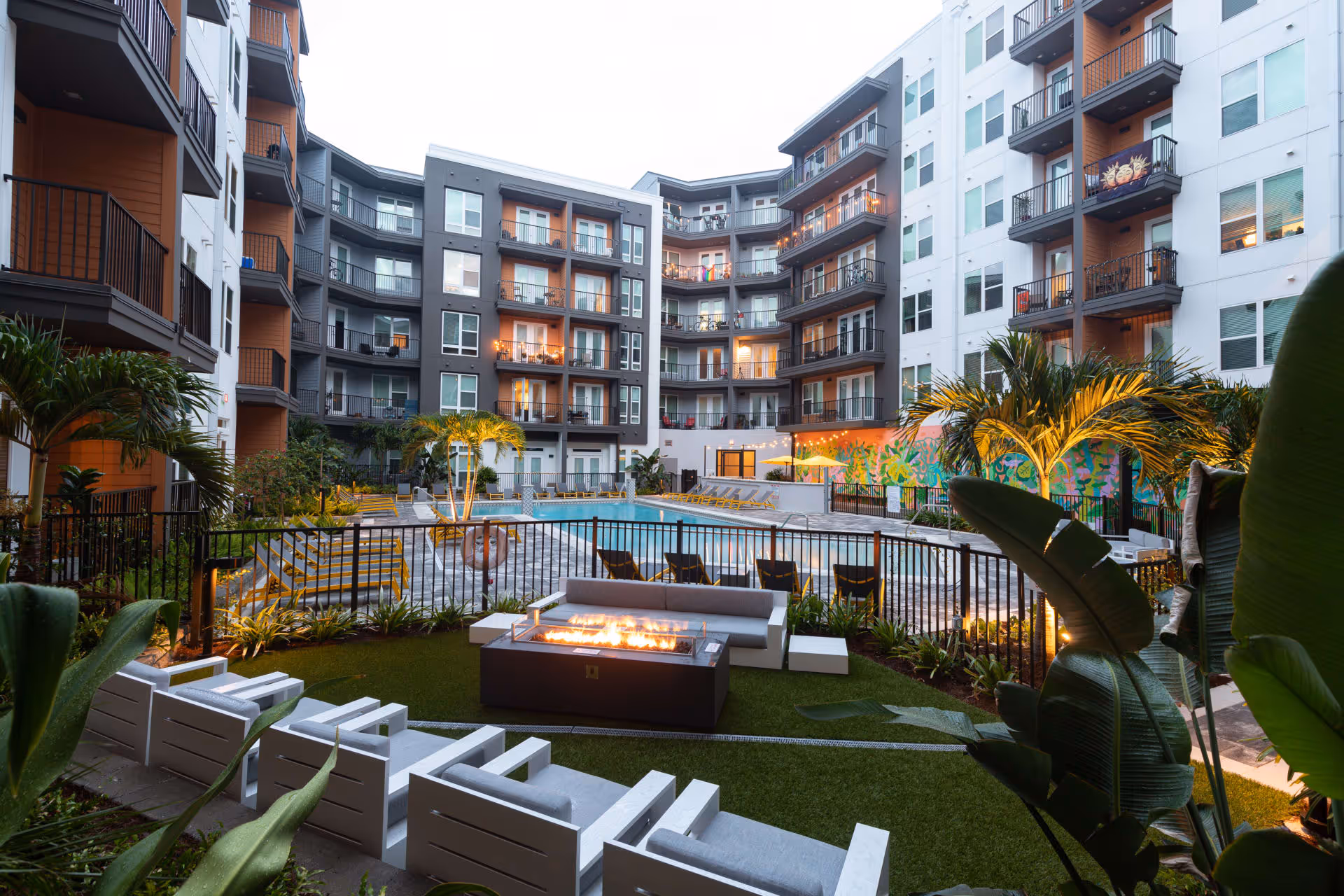 Outdoor courtyard area of a multi-story residential building with balconies, featuring a swimming pool surrounded by lounge chairs, a seating area with a fire pit, and tropical plants.