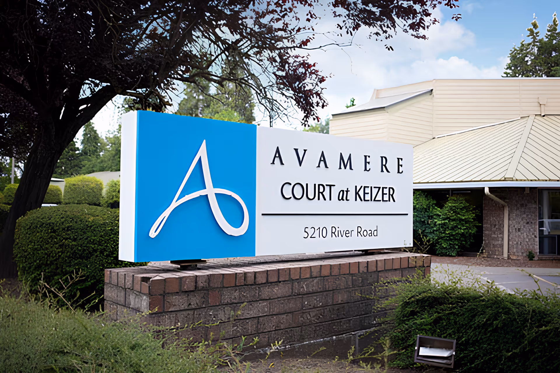 Outdoor sign reading "Avamere Court at Keizer, 5210 River Road" mounted on a brick base with the facility building and landscaping behind it.