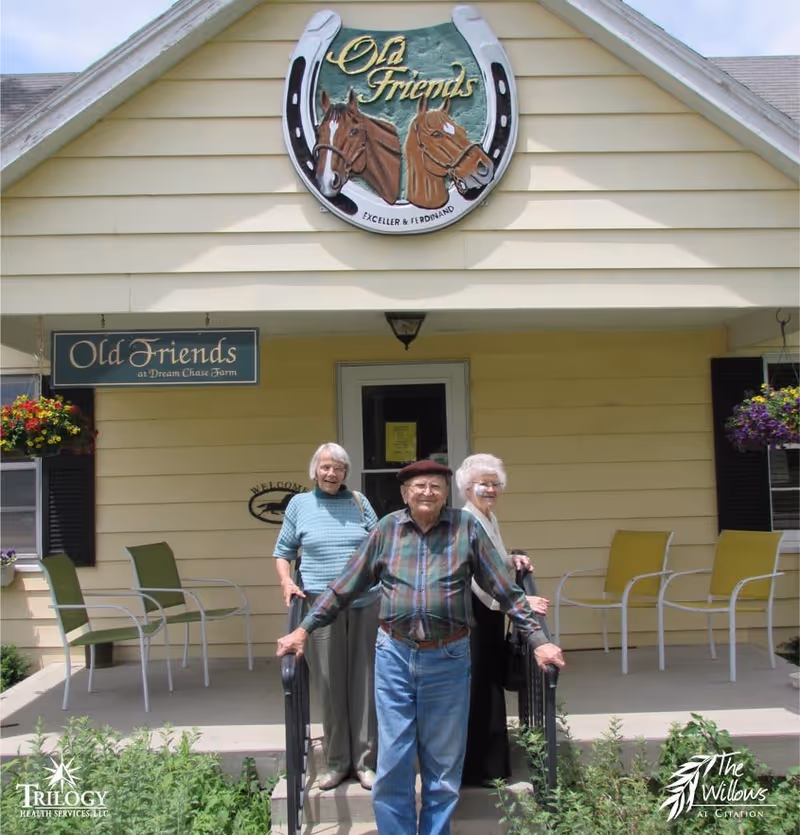 Three elderly people standing on the steps of a yellow building with a sign that reads 'Old Friends at Dream Chase Farm'. The building has a decorative sign above the entrance featuring two horses and the words 'Old Friends'. There are chairs on the porch and hanging flower baskets on either side.