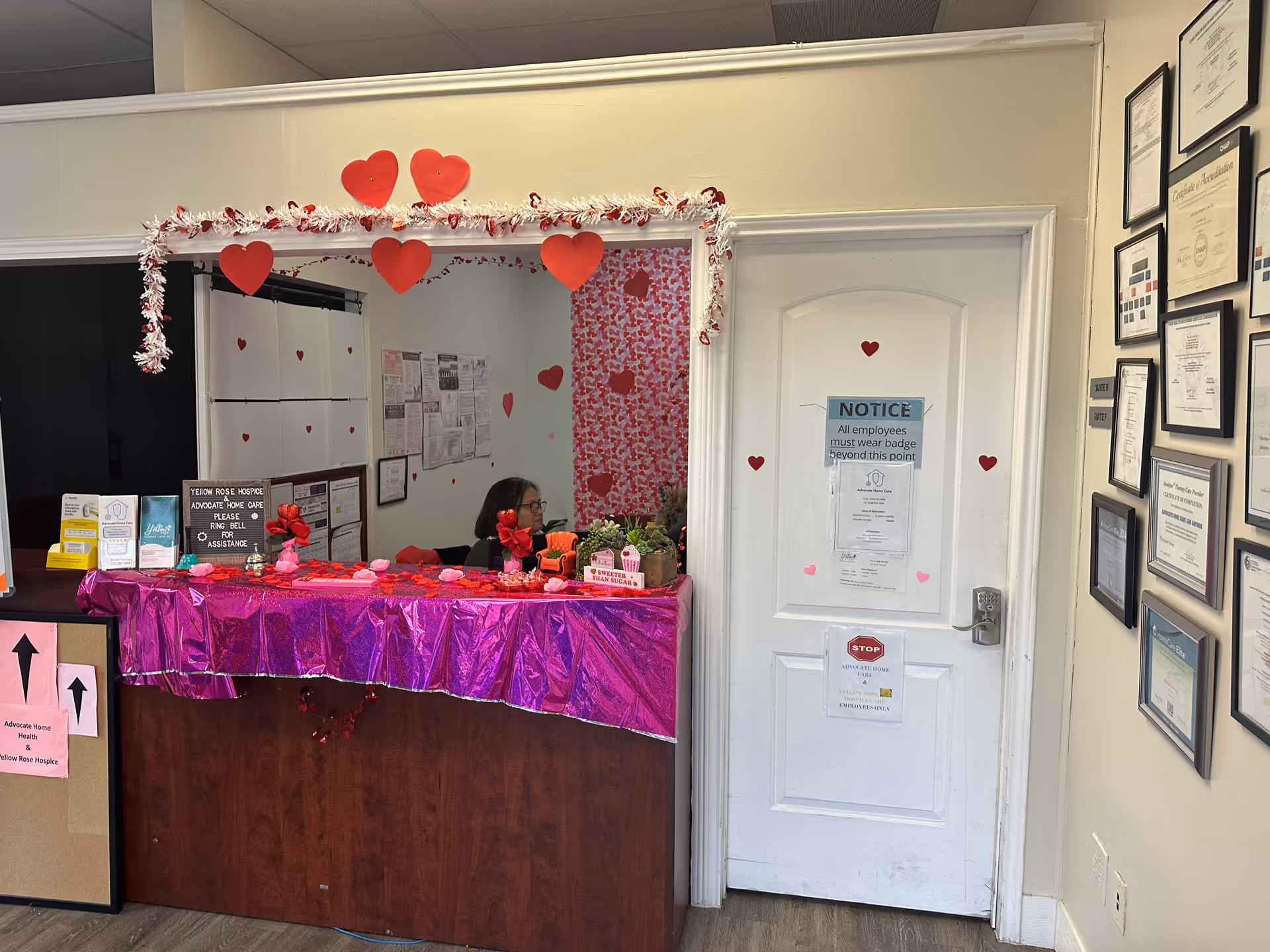 Reception desk in a Valentine-themed interior, with pink decorations, staff member, and framed certificates.