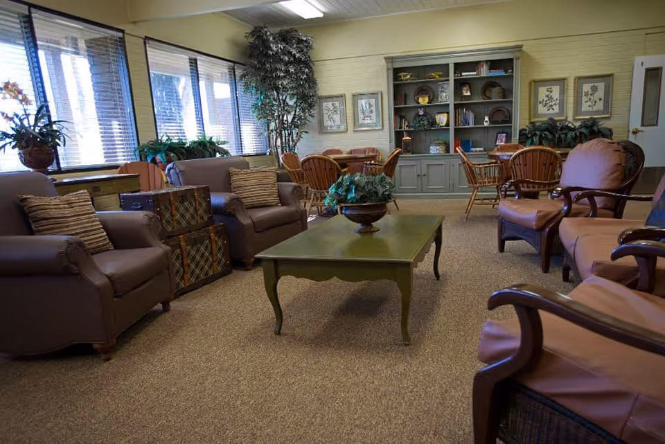 A cozy common room with armchairs and wooden chairs around a green coffee table, bookshelves and large windows along one wall.