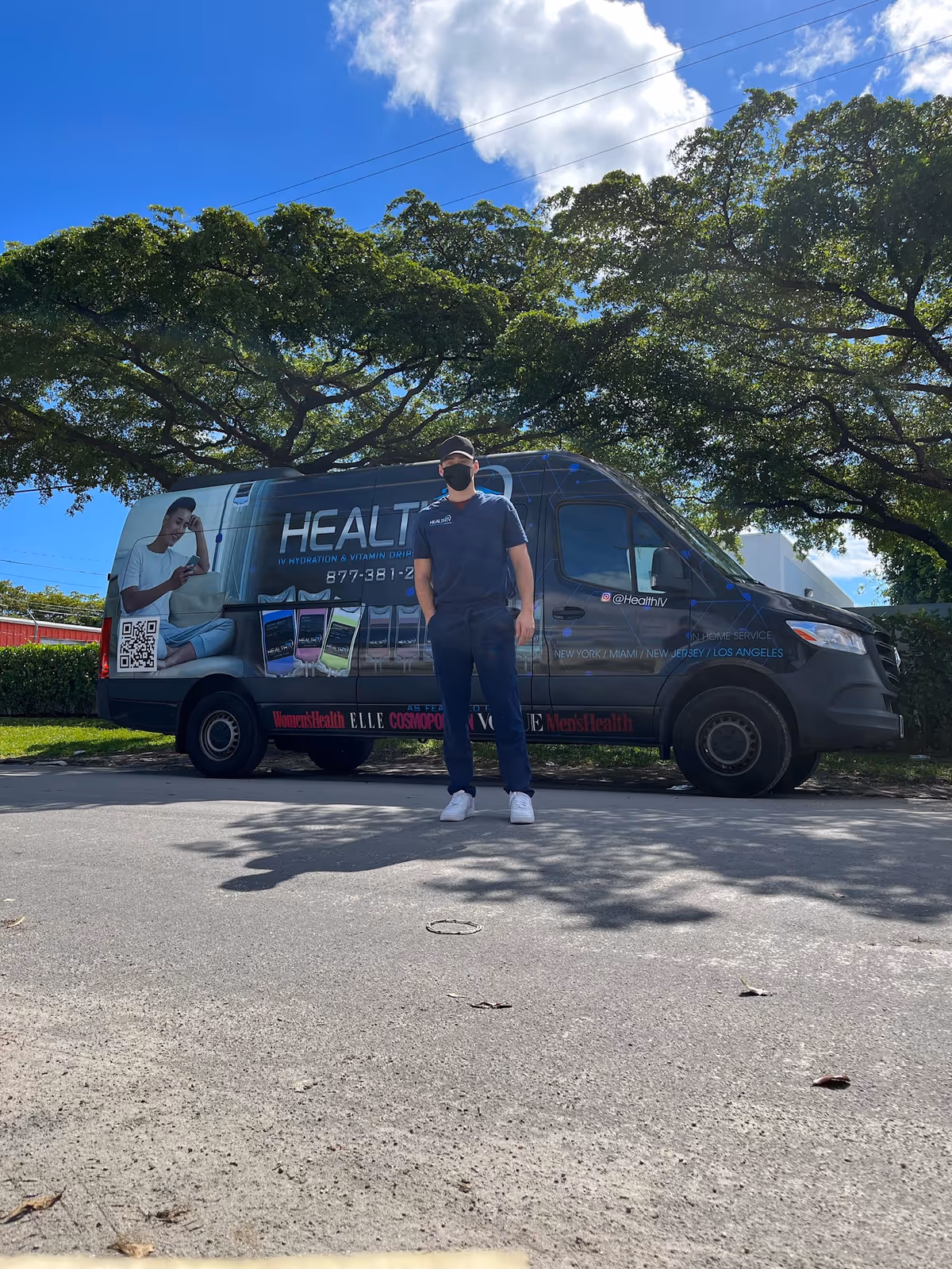 A man wearing a mask stands in front of a Health IV mobile therapy van outdoors.