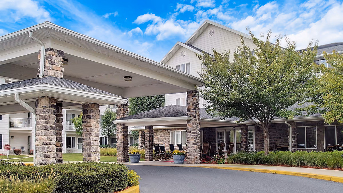Covered entrance of a senior living building with stone pillars, rocking chairs, and landscaped driveway.