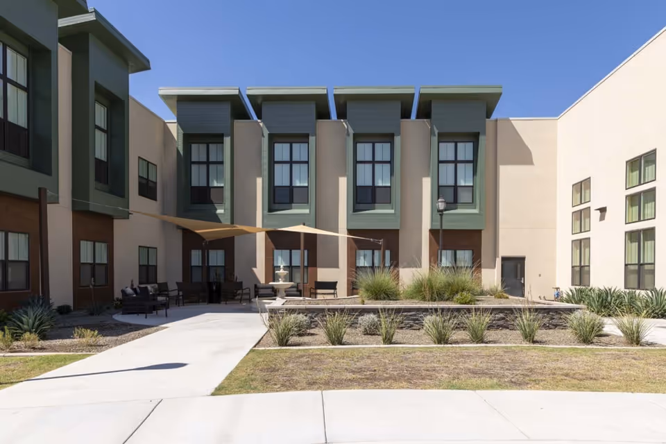 Courtyard with seating, a shaded canopy, fountain and landscaping in front of a two‑story rehabilitation center building.