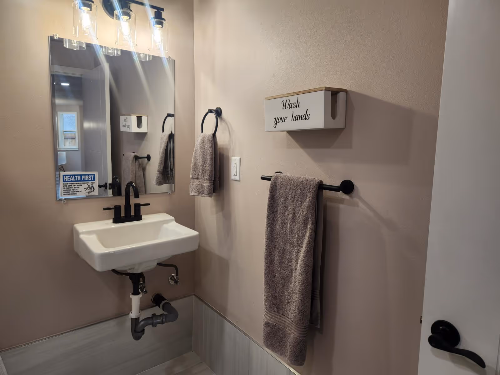 A bathroom sink area featuring a mirror, faucet, hand towels, and a sign that says 'Wash your hands.'