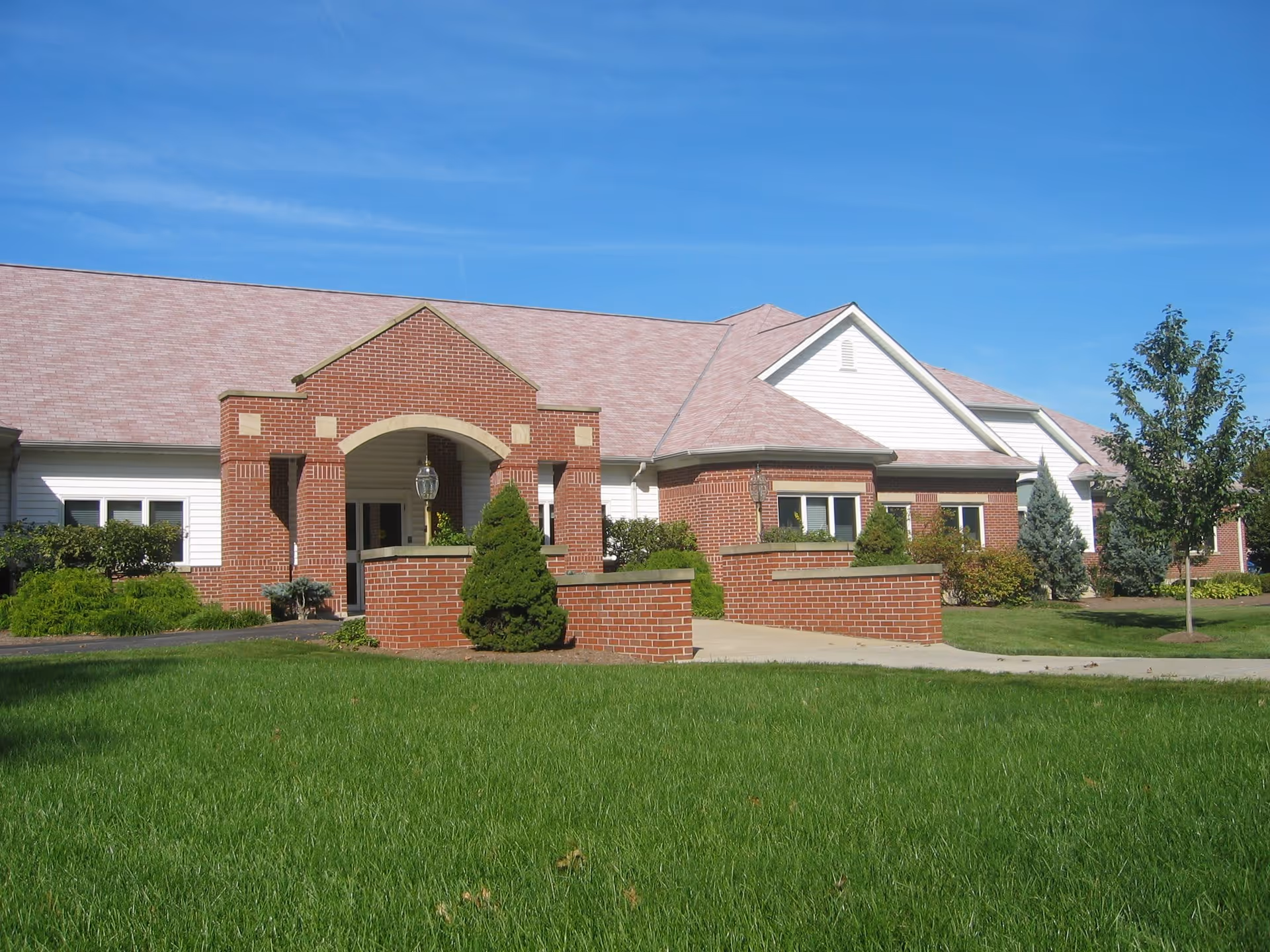 Exterior view of a single-story brick and white siding building with a covered entrance, surrounded by green grass, shrubs, and trees under a clear blue sky.