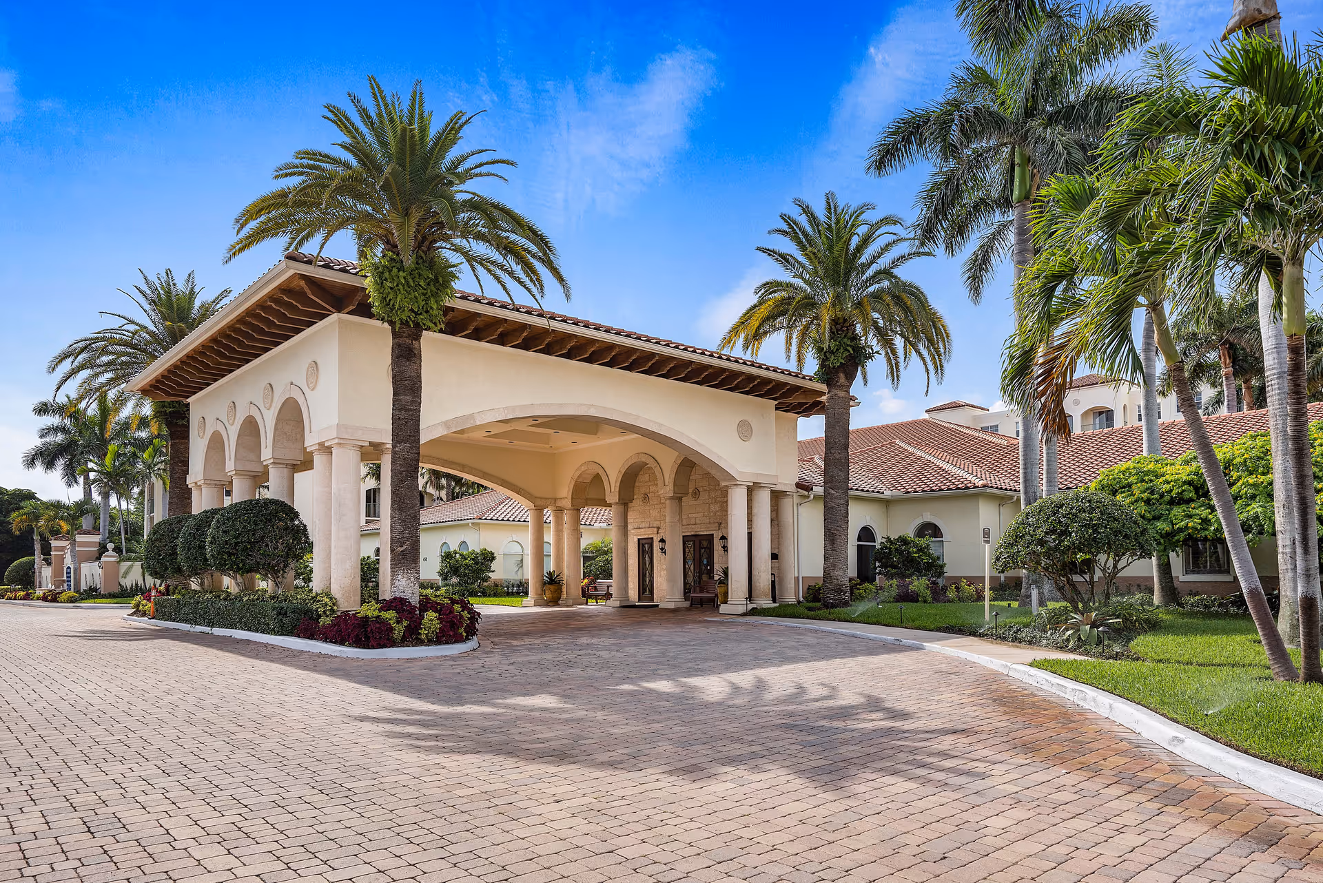 Entrance of The Residences at The Carlisle Palm Beach featuring a covered driveway with arches, surrounded by palm trees and well-maintained landscaping under a clear blue sky.