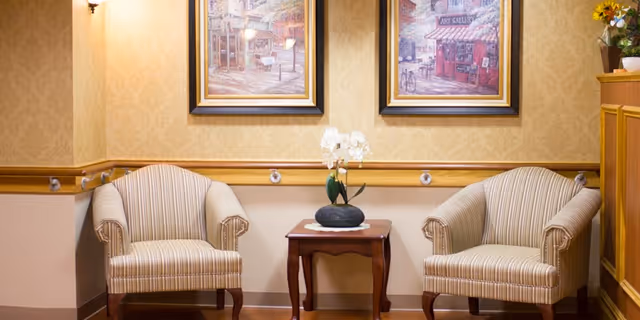 A cozy sitting area with two striped armchairs positioned on either side of a small wooden table. The table holds a decorative white orchid plant in a black pot. The wall behind features two framed paintings depicting quaint street scenes. The room has warm beige wallpaper and wooden trim with handrails along the wall.