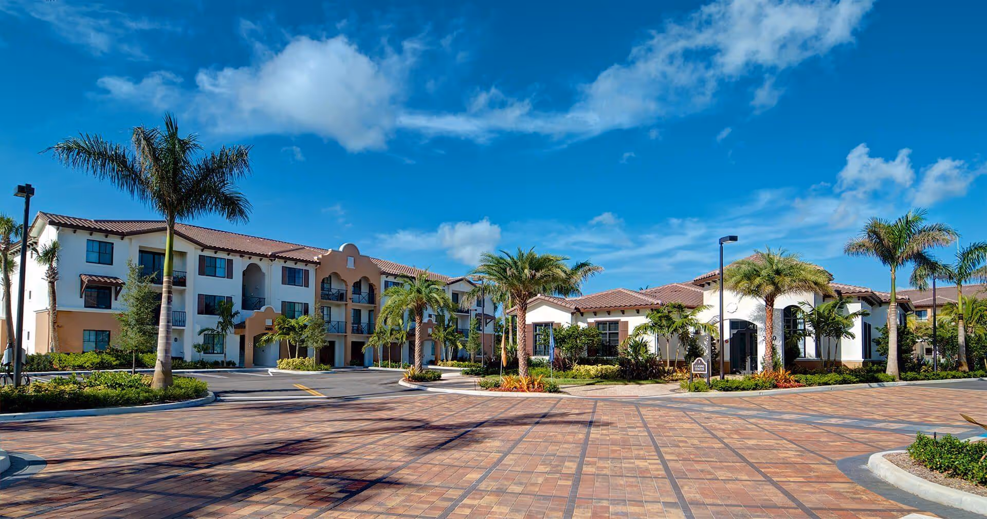 Exterior view of UpsideHoM Lighthouse Point facility showing a large paved driveway with palm trees and landscaped greenery. The building has a Mediterranean architectural style with white walls, terracotta roof tiles, and multiple windows. The sky is clear with some clouds.