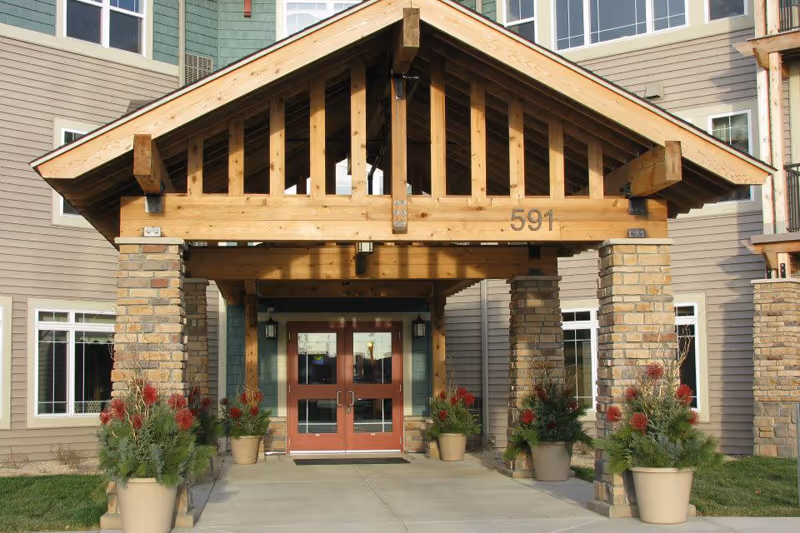 Covered wooden entrance with stone columns and double red doors at the front of a multi-story senior living building marked '591', flanked by potted plants.