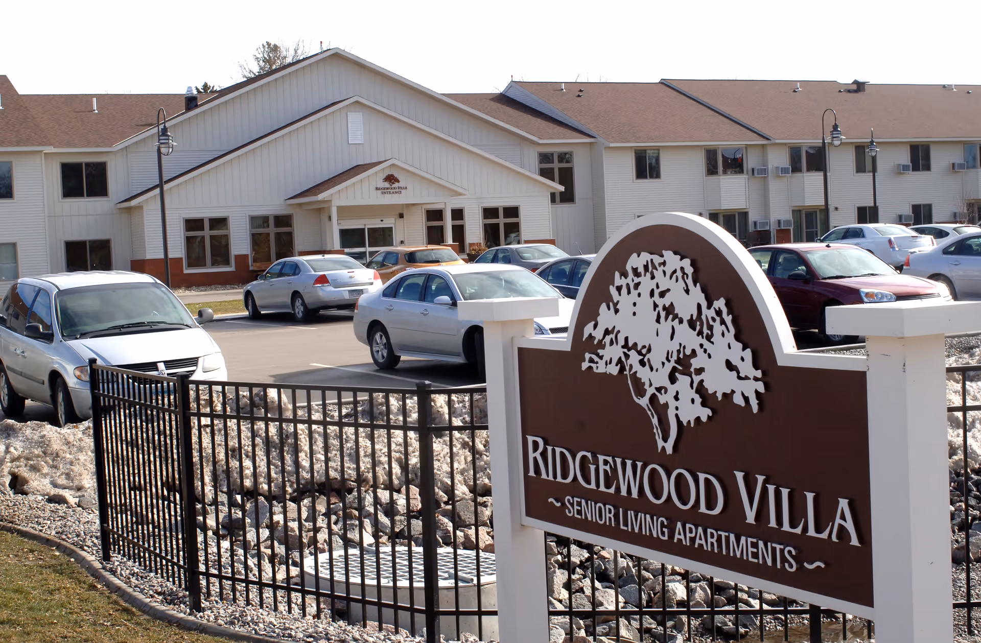 Exterior view of Ridgewood Villa Senior Living Apartments building with a parking lot in front. Several cars are parked, and a large brown and white sign with a tree logo reads 'Ridgewood Villa Senior Living Apartments'. The building is two stories with white siding and a brown roof.