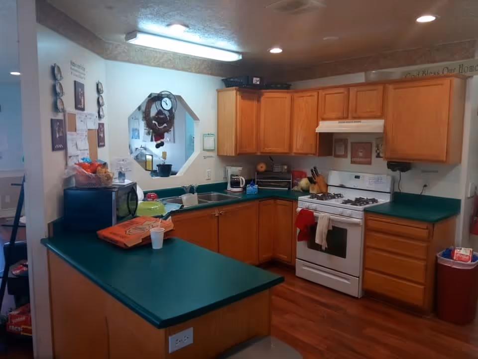 Interior view of a kitchen with wooden cabinets, green countertops, a white gas stove with oven, a microwave, a coffee maker, and various kitchen items including a pizza box and a cup on the counter. The kitchen has a window opening to another room and wooden flooring.