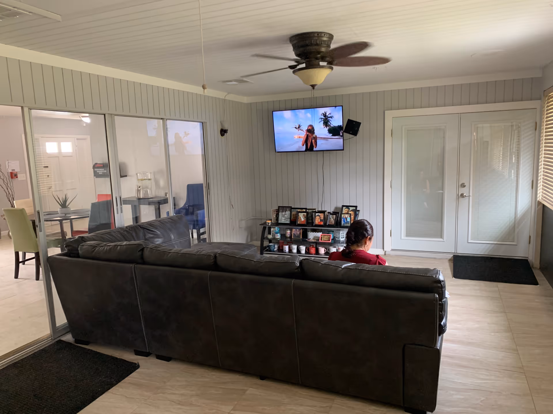 A living room area with a dark leather couch facing a wall-mounted TV displaying a person on a beach. A woman with braided hair is sitting on the couch. Below the TV is a shelf with framed photos and mugs. The room has light-colored walls with vertical paneling, a ceiling fan, and glass sliding doors leading to another room with tables and chairs.