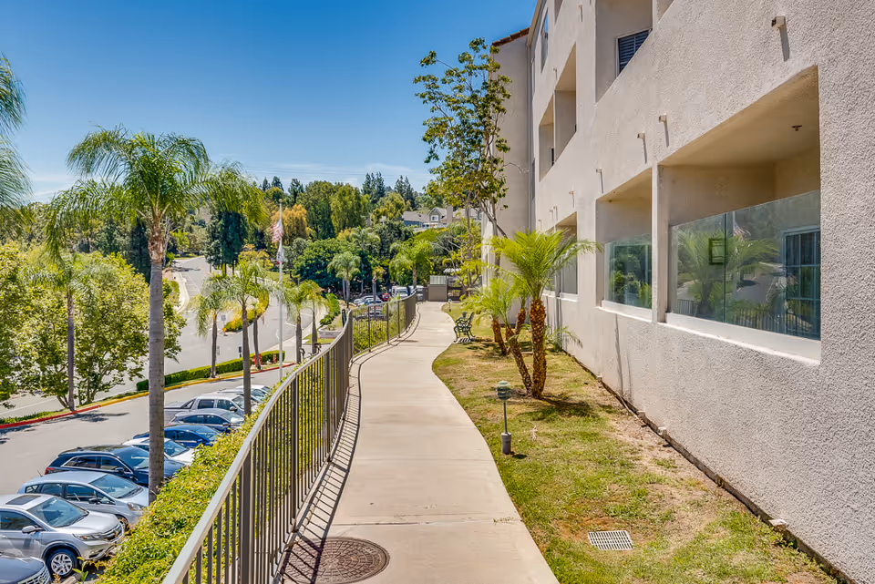 A sunlit exterior walkway beside a cream-colored senior living building with palm trees.