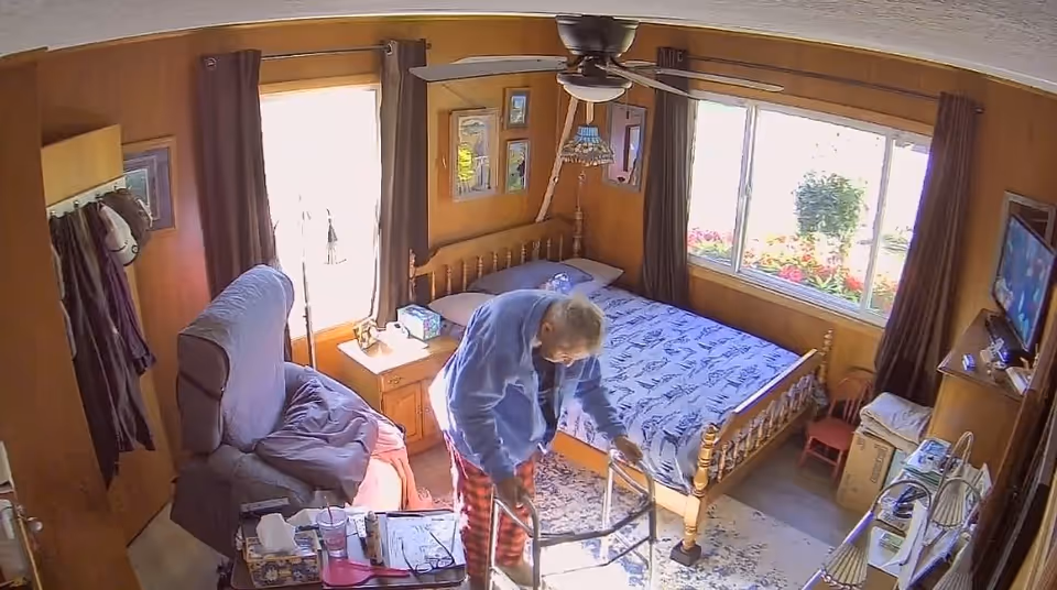 Senior resident in a blue shirt using a walker beside a bed in a cozy bedroom.