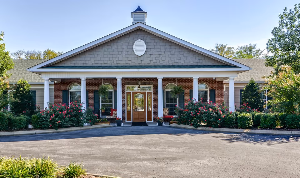 Front exterior view of a single-story brick building with a covered entrance supported by white columns. The entrance has a wooden door with glass panels and is flanked by windows with green shutters. There are hanging plants and flower bushes along the front, with a paved driveway in front of the building.