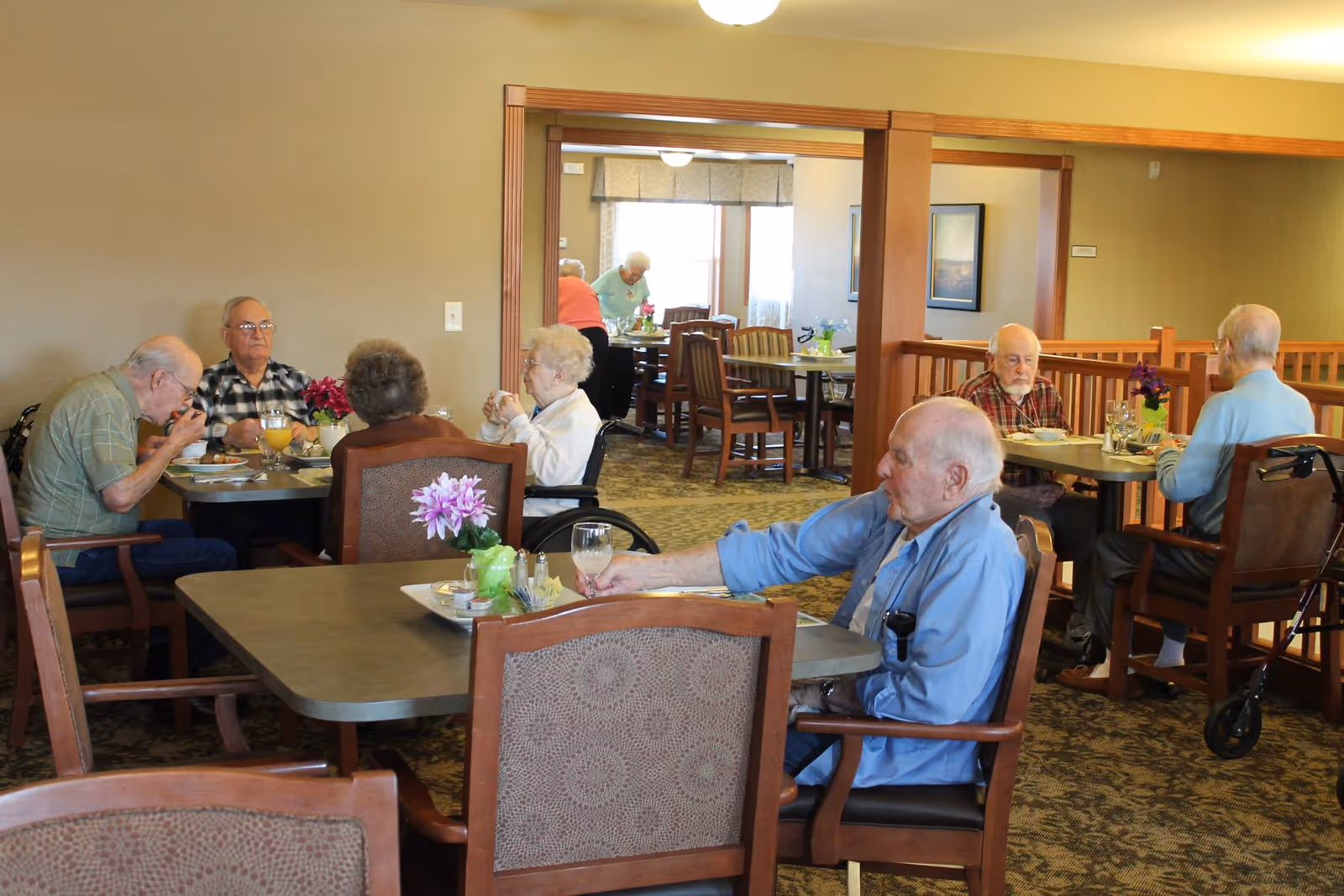 Elderly residents seated at tables eating and talking in a communal dining room with floral centerpieces.