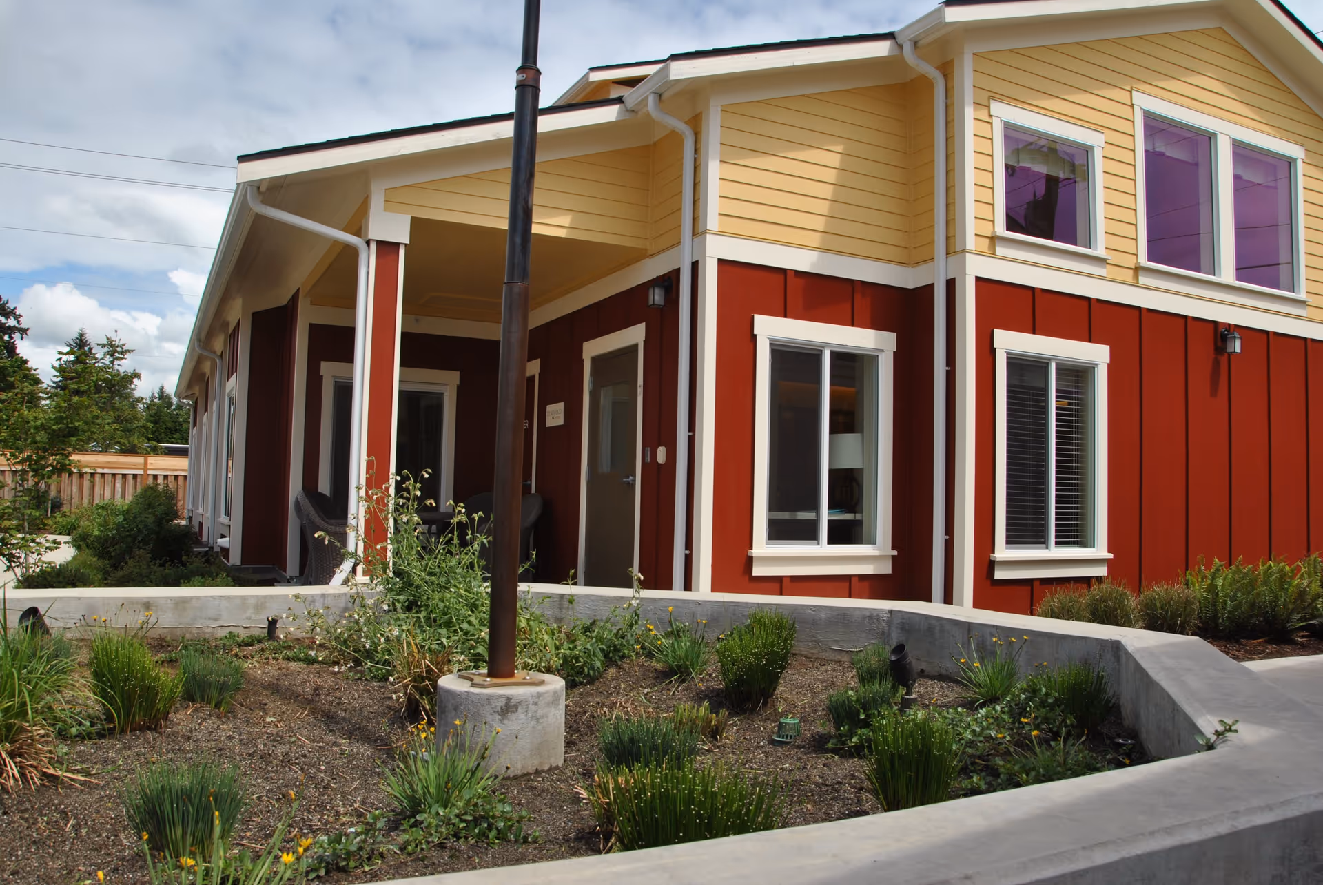 Exterior corner view of a red and yellow cottage building with windows, a covered porch entrance, and a landscaped planter bed.