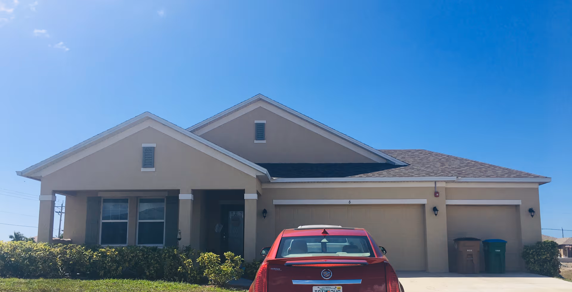 Front exterior view of a single-story beige house with a three-car garage and a red car parked in the driveway under a clear blue sky.
