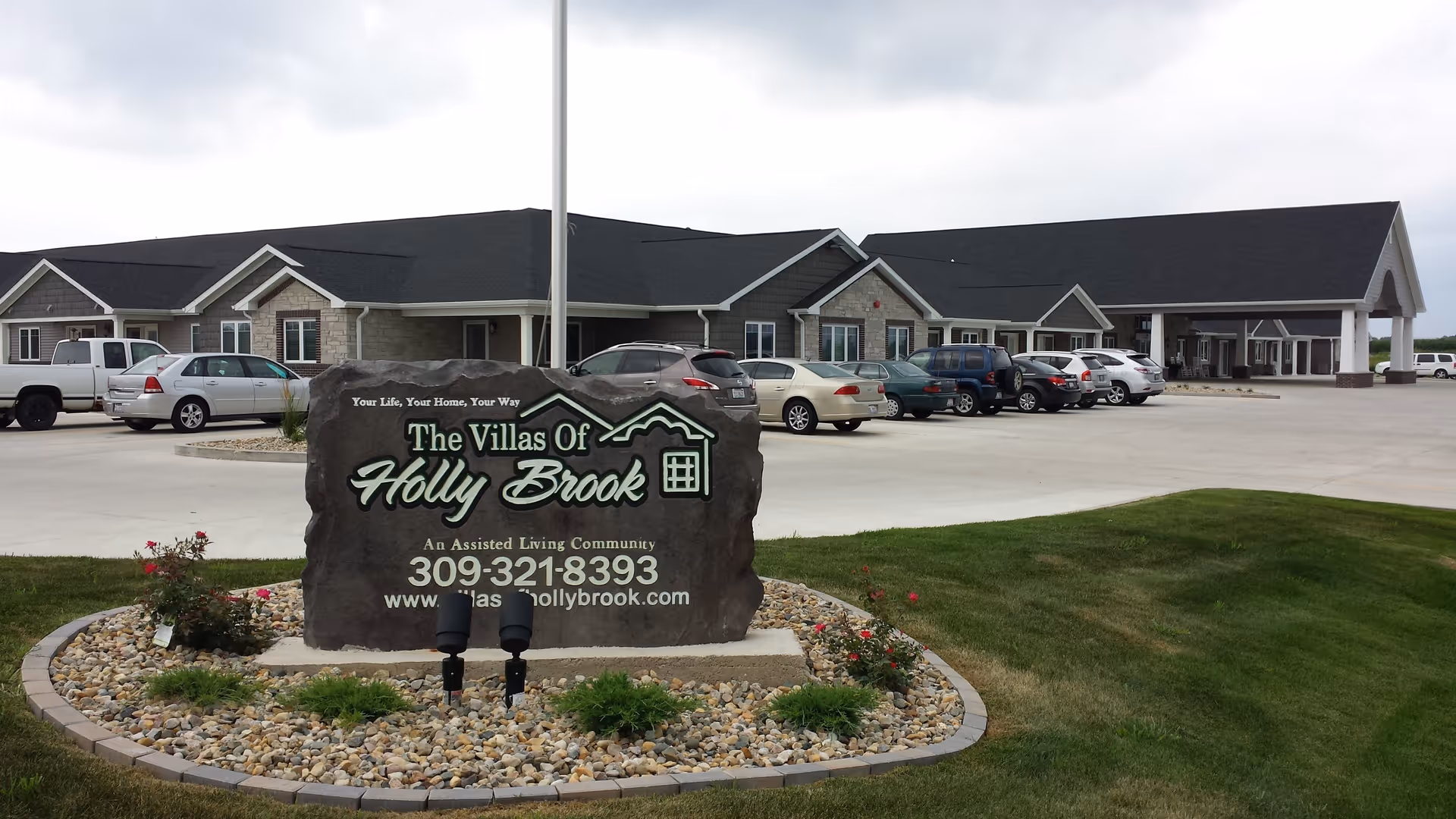 Exterior view of Villas of Holly Brook - Morton, showing a large stone sign with the facility's name, phone number, and website in front of a single-story building with a covered entrance and several parked cars.