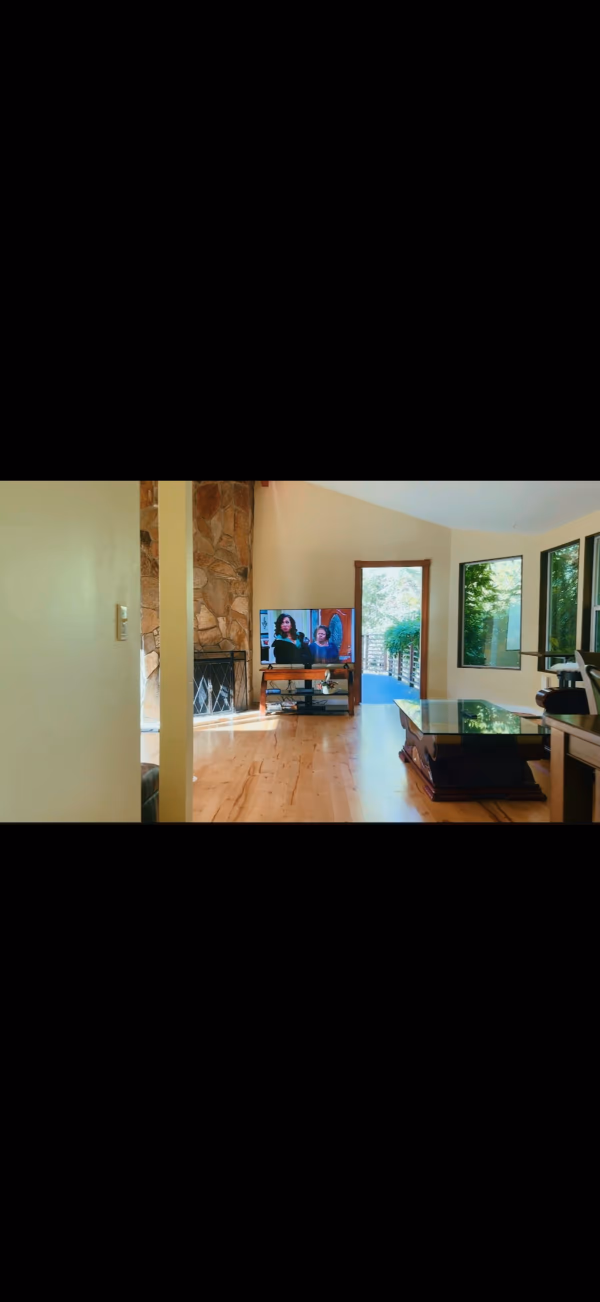 Bright living room with a stone fireplace, wooden floors, and a TV displaying people.