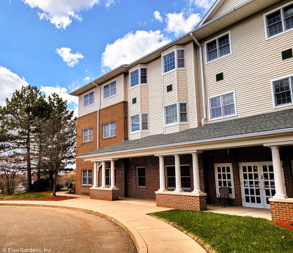 A multi-story senior living building exterior with brick base, beige siding, and a curved walkway.