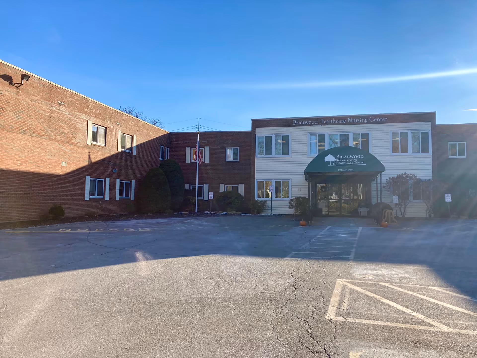 Exterior view of Briarwood Rehabilitation & Healthcare Center, a two-story building with a brick and white siding facade. The entrance has a green awning with the facility's name and address. There is a flagpole with an American flag in front, some bushes, and a clear blue sky above.