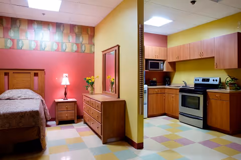 A room with a bed on the left side featuring a wooden headboard and a patterned bedspread. Next to the bed is a wooden nightstand with a lamp and a vase of yellow flowers. A wooden dresser with a mirror is positioned against the wall. On the right side, there is a kitchenette area with wooden cabinets, a microwave, a stove, and a sink. The floor has a colorful checkered pattern, and the walls are painted in warm tones of red and yellow.