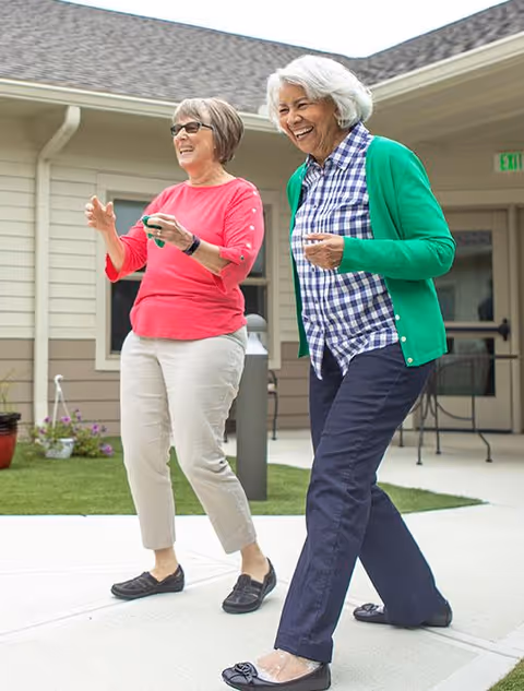 Two elderly women smiling and walking outdoors on a paved path near a building with beige siding and a gray roof. One woman is wearing a pink top and light-colored pants, while the other is wearing a green cardigan over a blue and white checkered shirt with dark pants. There are some potted plants and outdoor furniture visible in the background.