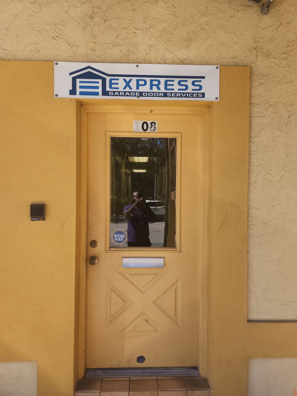 Yellow exterior door with Express Garage Door Services sign above, reflection of photographer in glass