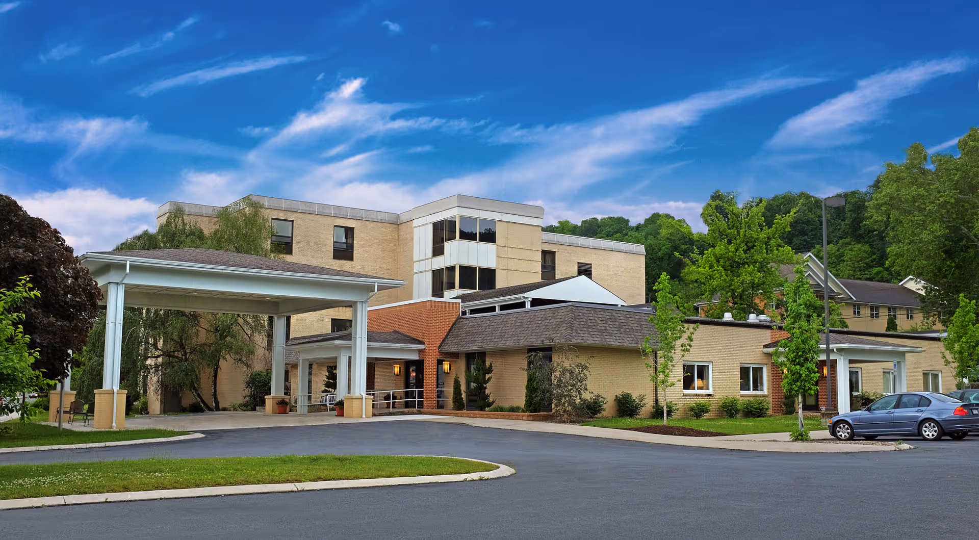 Exterior view of Epworth Manor Senior Living facility showing a multi-story beige brick building with a covered entrance driveway, surrounded by green trees and a clear blue sky.