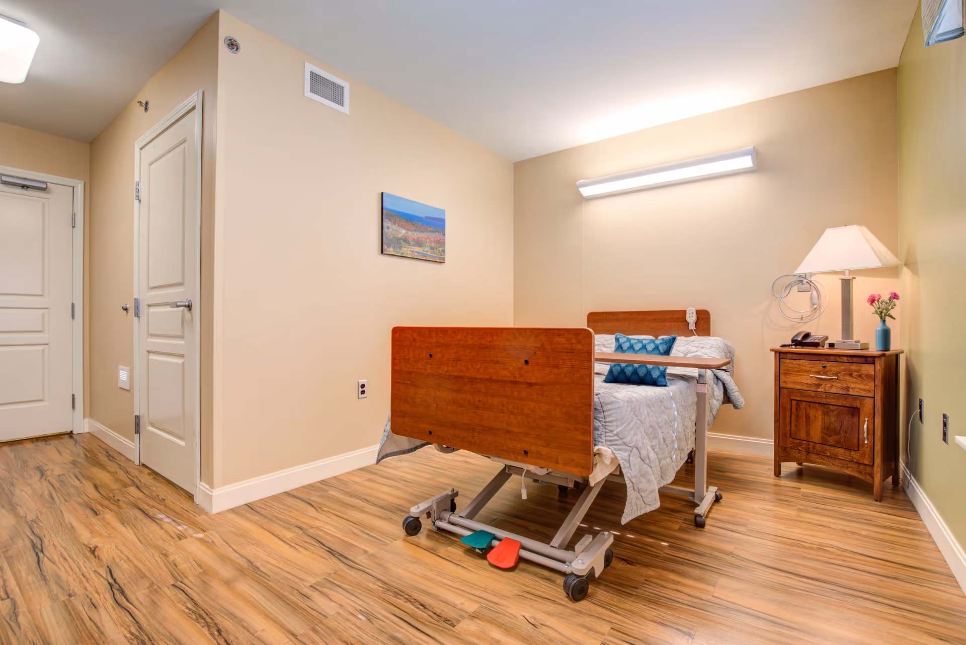 Hospital-style patient bed in a beige bedroom with a nightstand and lamp.