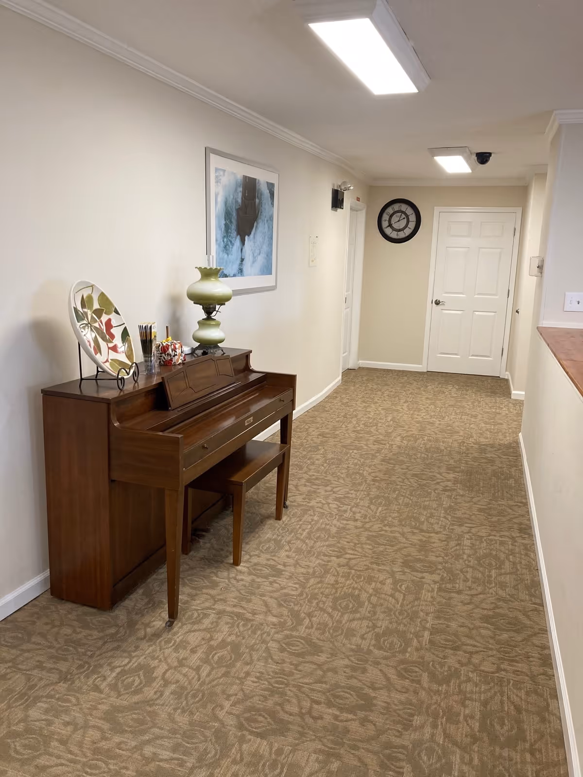 A hallway with beige patterned carpet and cream-colored walls. On the left side, there is a wooden piano with a matching bench, decorated with a large decorative plate, a green lamp, and some small items. Above the piano, a framed picture of a waterfall hangs on the wall. At the end of the hallway, there is a white door and a round wall clock. The ceiling has rectangular fluorescent lights and a security camera.