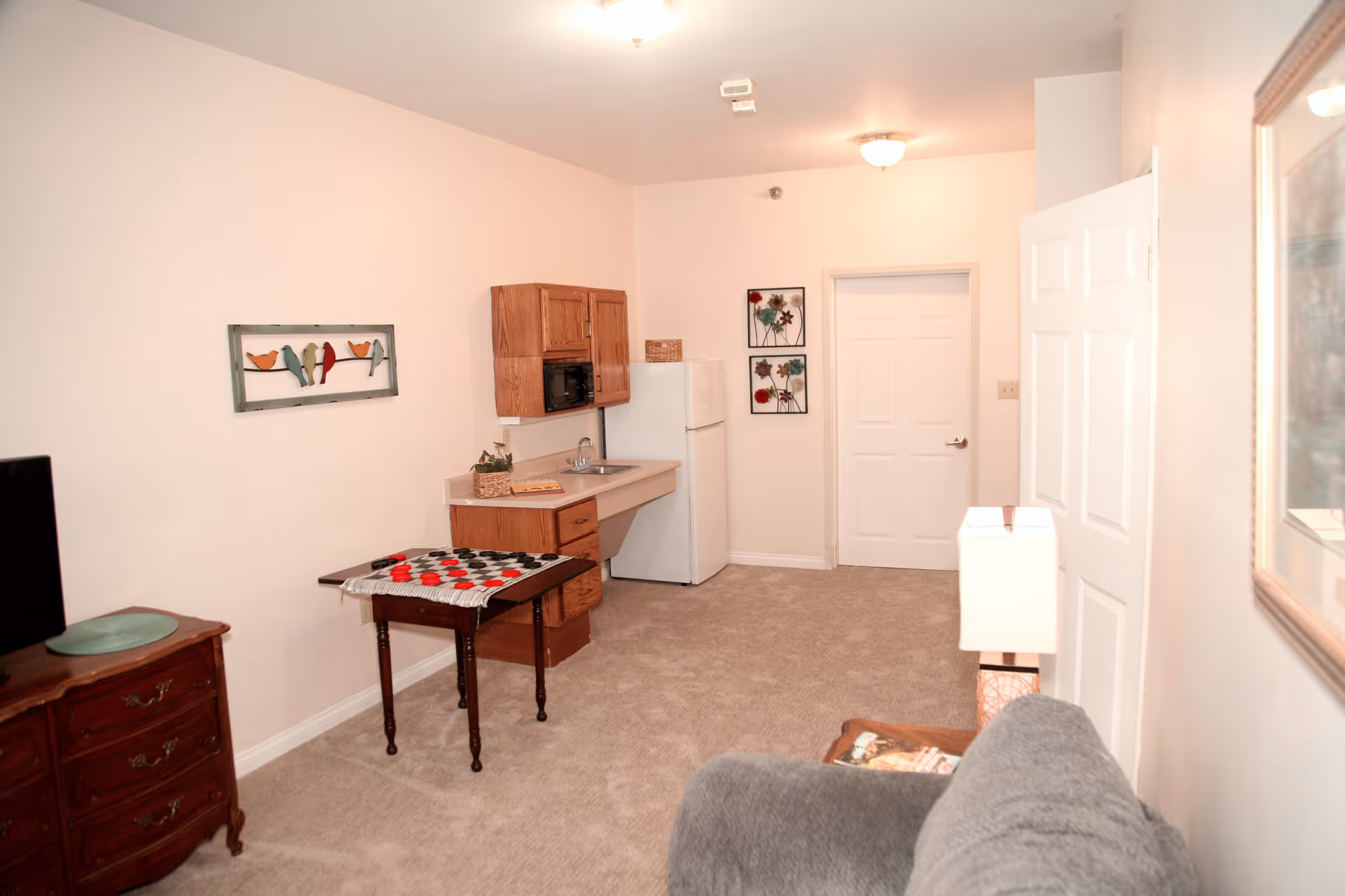 A small living space with beige walls and carpeted floor featuring a kitchenette with wooden cabinets, a white refrigerator, and a sink. There is a small table with a checkers board on it, a gray armchair, a wooden dresser with a TV on top, and wall decorations including a framed picture of birds and two floral artworks near a closed white door.
