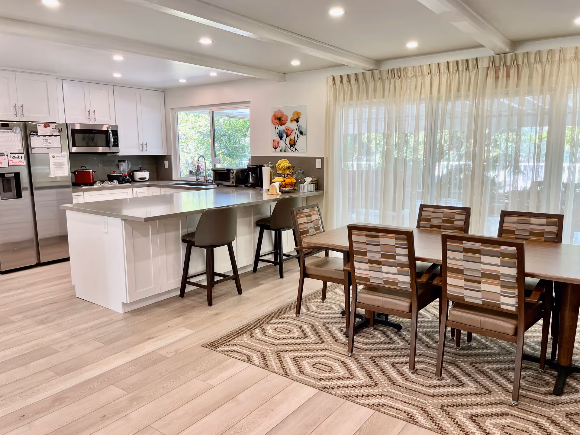 Bright open kitchen and dining area with island, stools, stainless appliances, and large windows.