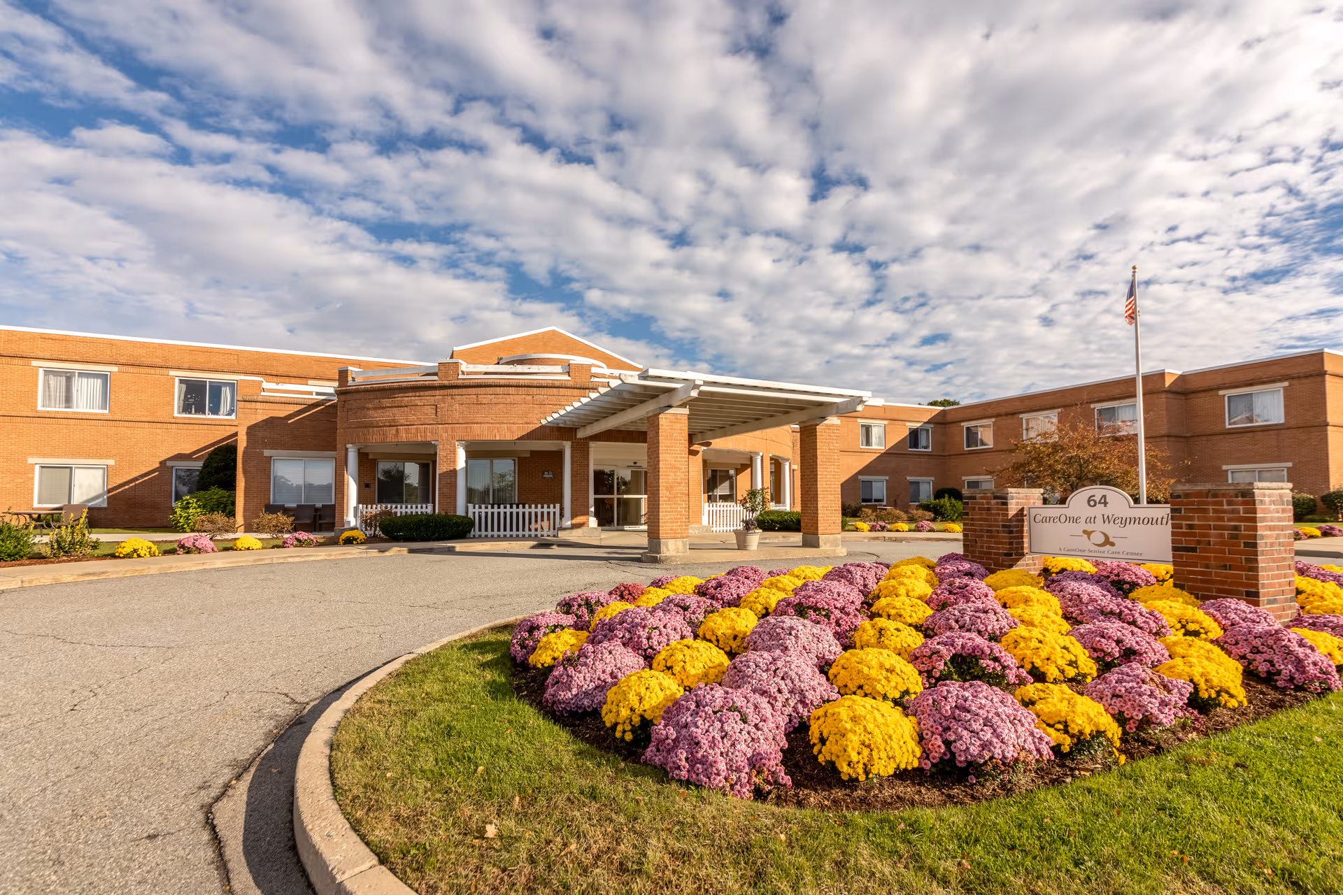 Exterior view of CareOne at Weymouth, a brick senior living facility with a covered entrance and a circular driveway. In front of the building is a landscaped area with neatly arranged yellow and purple flowers and a sign displaying the facility's name and address number 64. The sky is partly cloudy.